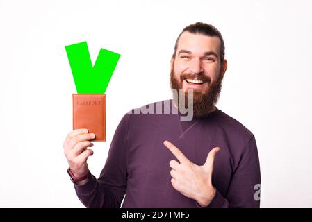 Photo d'un homme joyeux barbu qui tient un passeport avec quelques billets dans lui pointant vers eux est de regarder la caméra . Banque D'Images