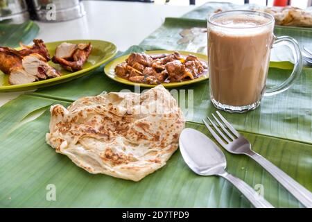 Roti canai ou paratha servi sur une feuille de banane, avec du curry de mouton et du poulet frit, et du thé populaire tarik Banque D'Images