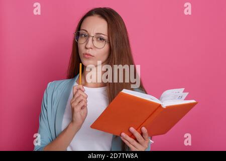 Image d'une femme intelligente et réfléchie portant des lunettes, tenant un carnet orange et un stylo, en planifiant son temps, en utilisant un organiseur, debout isolé sur rose Banque D'Images