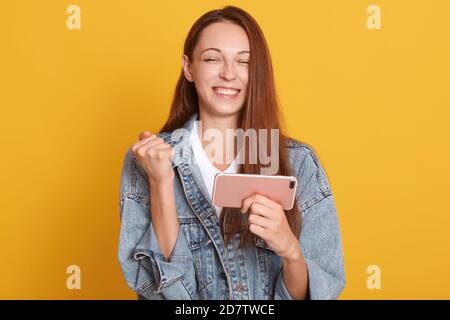 Photo d'une jeune femme heureuse avec de beaux cheveux longs accrochant poing tout en tenant le téléphone portable dans la main, portant l'élégante veste demin, posant avec près Banque D'Images