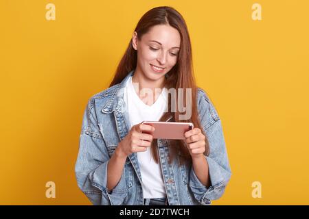 Image d'une fille attrayante et tendre souriant sincèrement, en tenant le smartphone rose dans ses mains, en regardant son écran, en regardant la vidéo, en étant affectueux de la nouvelle technologie Banque D'Images