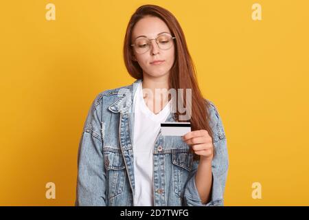 Jeune belle femme caucasienne présentant la carte de crédit pour faire le paiement, adorable dame regarde la carte, portant la veste en denim et t-shirt blanc, pense Banque D'Images