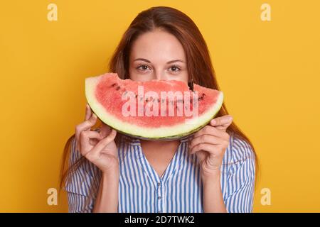 Image d'une jeune adorable femme habillée d'une chemise rayée élégante, tenant un morceau de pastèque dans les mains, une femme se cachant derrière le mamelon d'eau et regardant directement Banque D'Images