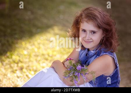 Une jeune fille de 9 à 10 ans avec un bouquet de fleurs sauvages bleues est assise sur une pelouse verte dans le parc. Une promenade dans le parc en été.