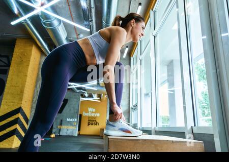 Jeune belle femme de fitness dans les vêtements de sport nouant des lacets et regardant la fenêtre tout en faisant de l'exercice à la salle de gym. Sport, bien-être et mode de vie sain Banque D'Images