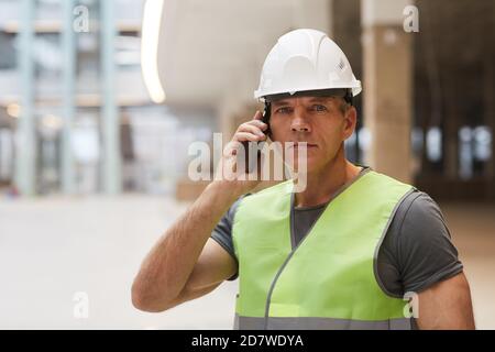 Portrait d'un travailleur de la construction professionnel parlant au téléphone et regardant l'appareil photo tout en se tenant sur le chantier, espace de copie Banque D'Images