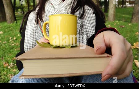 Une fille vêtue d'un manteau et d'un foulard dans la forêt d'automne tient un livre et une tasse avec une boisson chaude dans ses mains en gros plan dans un parc de la ville lors d'une journée chaude. Banque D'Images