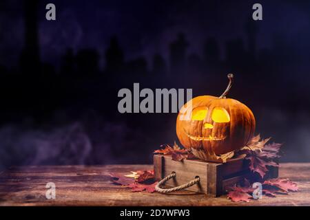 Halloween concept avec colère et triste lampe de citrouille avec des feuilles d'automne sèches et colorées sur l'ancienne table en bois avec un cimetière sombre sur le fond. Banque D'Images