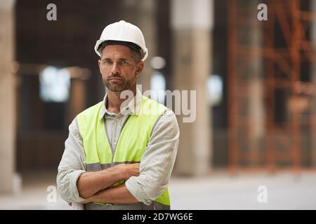 Portrait à la taille de l'ouvrier de construction mature regardant la caméra tout en se tenant debout avec les bras croisés sur le chantier de construction, l'espace de copie Banque D'Images