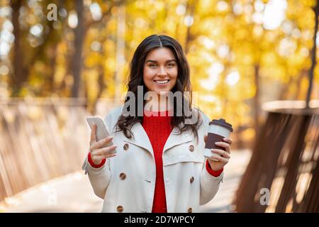 Portrait d'une jolie dame avec café à emporter et smartphone souriant et regardant l'appareil photo à l'extérieur par beau temps d'automne Banque D'Images