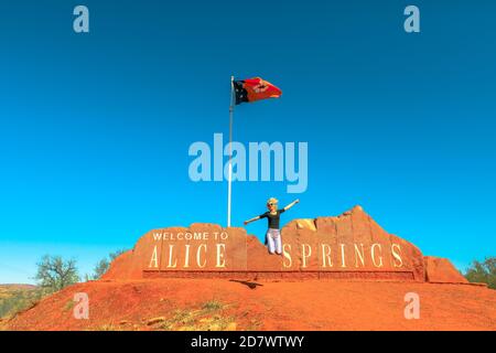 Femme touristique insouciante sautant à Alice Springs Bienvenue panneau dans le territoire du Nord, Australie centrale. Tourisme dans le désert de l'Outback Red Centre. Déplacement Banque D'Images