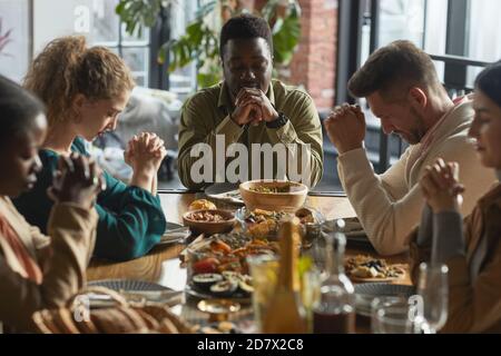 Portrait de l'homme afro-américain priant avec les yeux fermés tout en étant assis à la table du dîner pendant la célébration de Thanksgiving avec des amis et la famille, copier l'espace Banque D'Images