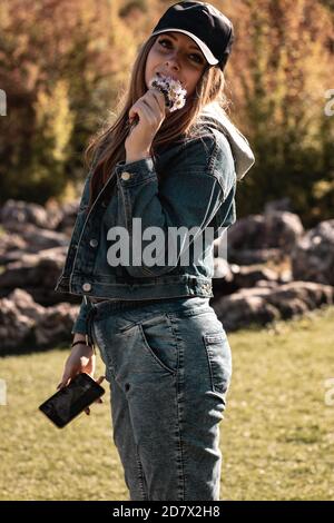 Gros plan vertical d'une jolie brune portant un Jean bleu et une veste, tenant un smartphone et un bouquet de fleurs dans une main, souriant et enjo Banque D'Images