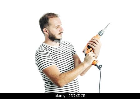Un homme barbu dans un T-shirt rayé, tient une perceuse électrique dans ses mains avec un look de questionnement. Banque D'Images