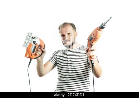 Un homme sérieux dans un T-shirt rayé, tient une perceuse et une scie électrique dans ses mains. Isolé sur fond blanc Banque D'Images