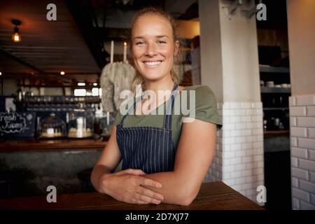 Jeune serveuse gaie vêtue d'un tablier debout et assise sur une table dans le café Banque D'Images