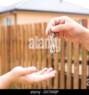 Main avec une clé et un porte-clés en bois. Arrière-plan de la clôture et du chalet. Construction, projet, déménagement à une nouvelle maison, hypothèque, location et achat rea Banque D'Images