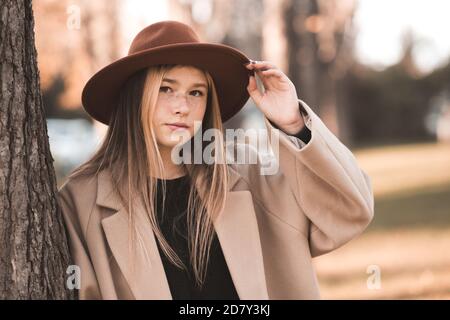 Adolescente souriante de 13-14 ans portant un chapeau élégant et un manteau d'hiver beige posant sur un arrière-plan d'automne gros plan. Teenagerhood. Banque D'Images