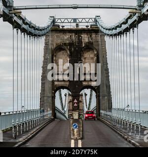 Une voiture rouge passe sous les arches du pont suspendu historique de Menai, pays de Galles conçu par Thomas Telford Banque D'Images