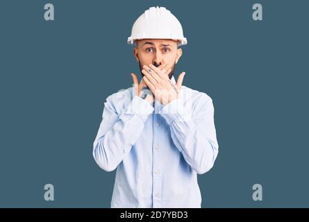 Jeune homme beau portant un casque d'architecte choqué couvrant la bouche avec des mains pour erreur. Concept secret. Banque D'Images