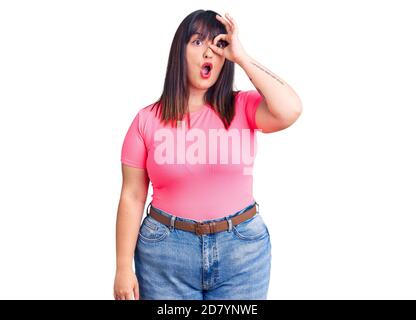 Jeune femme de taille plus portant des vêtements décontractés faisant geste ok choqué avec le visage surpris, l'oeil regardant à travers les doigts. Expression incroyant. Banque D'Images