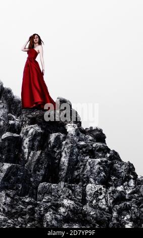 Belle femme aux cheveux rouges debout sur une colline rocheuse dans un rouge robe Banque D'Images