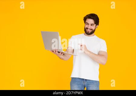 Portrait d'un jeune homme barbu souriant tenant un ordinateur portable ordinateur tout en regardant et pointant sur l'écran dans un studio isolé arrière-plan jaune Banque D'Images