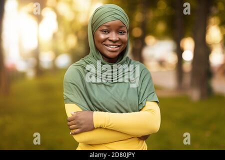 Femme musulmane noire souriante dans le hijab debout avec les bras pliés À l'extérieur Banque D'Images