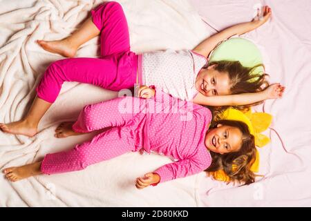 Les enfants aux visages souriants sont sur fond de couverture rose clair. Les écolières ont une fête de pyjama, vue du dessus. Enfants en pyjama à pois roses avec les mains levées. Concept d'enfance et de bonheur. Banque D'Images