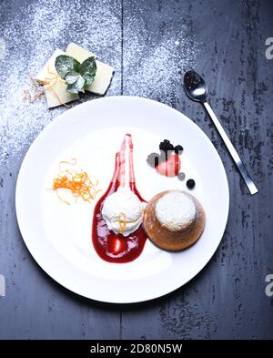 Dessert servi avec des nouilles orange dans un café ou une boulangerie. Gâteau de lave fondu avec baies sur l'assiette. Fondant au chocolat blanc avec glace à la vanille et sauce aux fraises sur fond gris. Concept de pâtisserie Banque D'Images