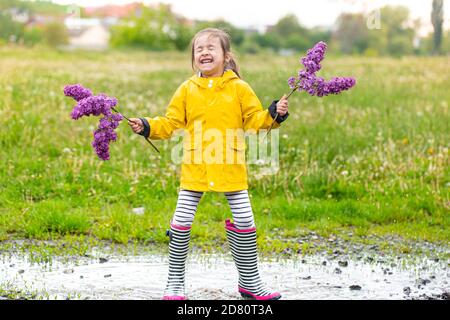 Une petite fille gaie et mignonne en bottes de caoutchouc tient dans un flaque et tient des fleurs de lilas dans ses mains. Banque D'Images