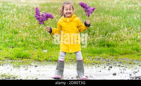 Une petite fille gaie et mignonne en bottes de caoutchouc tient dans un flaque et tient des fleurs de lilas dans ses mains. Banque D'Images