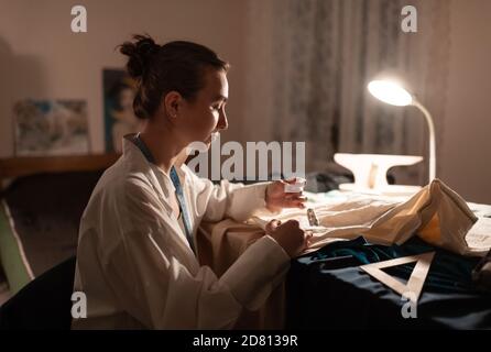 Vue latérale de calme femme tailleur assis à la table avec lampe à brûler et pièces de tissu à coudre Banque D'Images