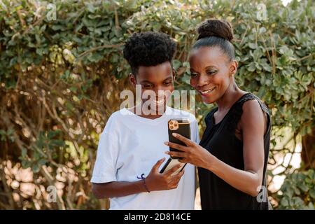 Une maman noire souriante et un fils préadolescent regardant les téléphones portables ensemble Banque D'Images