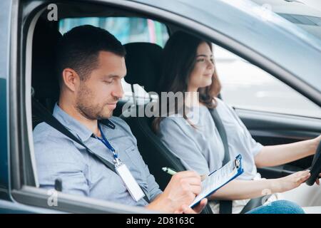 Apprendre à conduire une voiture. Un instructeur et une étudiante de sexe masculin s'exerçant à conduire. Le jeune homme remarque les résultats du test dans son article. Femme gaie satis Banque D'Images