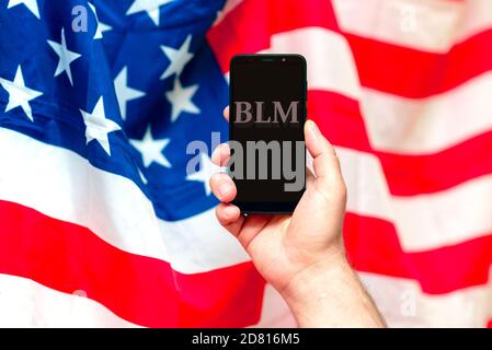 Main de l'homme tenant un téléphone portable avec l'abréviation de texte de Black Lives importe contre le drapeau flou des États-Unis. Banque D'Images