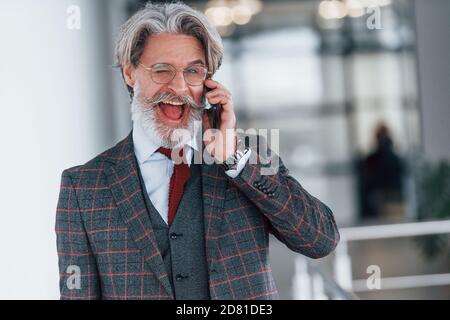 Homme d'affaires senior en costume et cravate avec cheveux gris et barbe debout à l'intérieur avec téléphone Banque D'Images