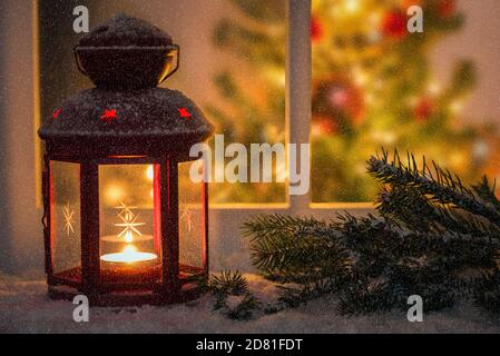 Lanterne rouge allumée sur un rebord extérieur recouvert de neige pendant une nuit de neige. Un arbre de Noël à l'intérieur de la maison dans visible par la fenêtre. Banque D'Images