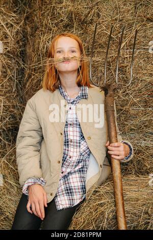 Petite fille de gingembre drôle faisant la moustache avec les lames de foin à l'intérieur une grange Banque D'Images