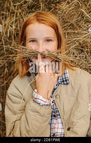 Portrait d'une petite fille de gingembre qui fait de la moustache avec du foin lames à l'intérieur d'une grange Banque D'Images