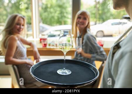 un serveur inreconnaissable au café a apporté des verres de champagne sur le plateau aux jeunes clientes. les femmes blondes attrayantes s'assoient en regardant un serveur agréable et agréable. gros plan sur le verre Banque D'Images