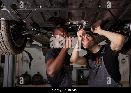 deux mécaniciens interraciaux divers vérifiant le fond de la voiture en service auto, coopérer, travailler ensemble Banque D'Images