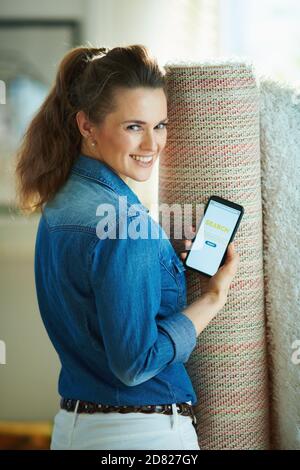 femme moderne souriante en jean et pantalon blanc avec tapis blanc à la recherche d'un service de nettoyage de tapis sur un smartphone à la maison moderne dans da ensoleillé Banque D'Images