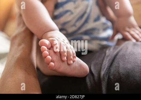 Foyer sélectif des mains touchant les jambes et les pieds du bébé garçon en se reposant et assis sur le ventre de son père avec assistance à domicile Banque D'Images