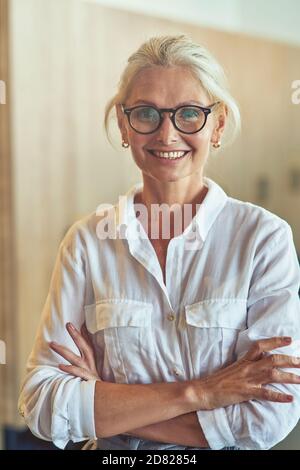 Photo verticale d'une jeune femme d'affaires mûre et heureuse chemise blanche regardant l'appareil photo et souriant tout en posant à l'intérieur le bureau Banque D'Images