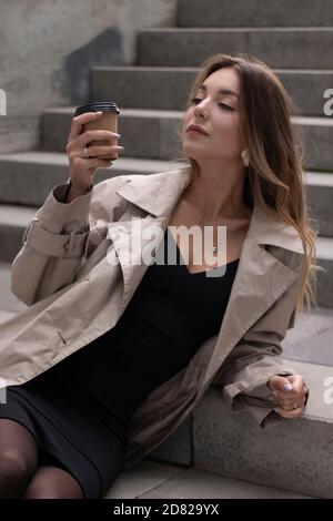 jolie petite brunette mince dans une tranchée à la mode et robe noire assise sur un escalier en béton gris avec une tasse de café en papier brun. Banque D'Images