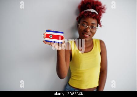 Femme afro-américaine à cheveux afro, porter un maillot jaune et des lunettes, tenir le drapeau Costa Rica isolé sur fond blanc. Banque D'Images