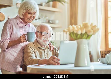 Souriante belle femme âgée tenant une tasse de café et se tenant près de son mari tout en regardant la vidéo sur ordinateur. Concept de style de vie domestique Banque D'Images