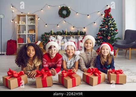 Groupe d'enfants divers qui se trouvent sur le sol près des cadeaux de Noël et qui regardent l'appareil photo. Banque D'Images