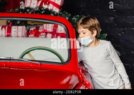 Mignon petit garçon sérieux dans les masques médicaux se préparent pour la nouvelle année près de la voiture de Noël. Vacances, distance sociale, rester à la maison. Banque D'Images